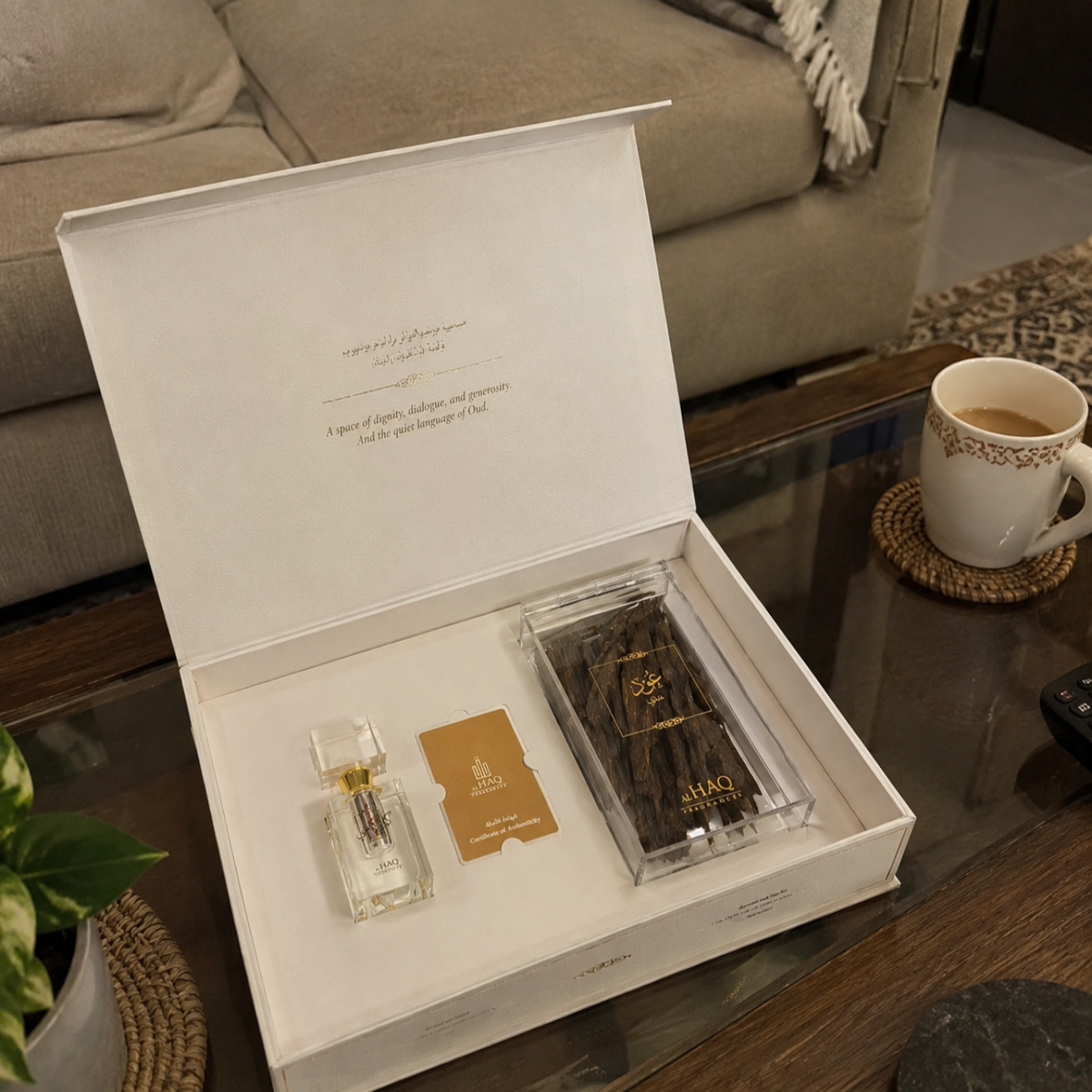 An open white gift box displayed on a glass coffee table in a living room setting, containing a small clear perfume bottle with gold detailing and a clear case holding dark brown incense sticks with gold branding.